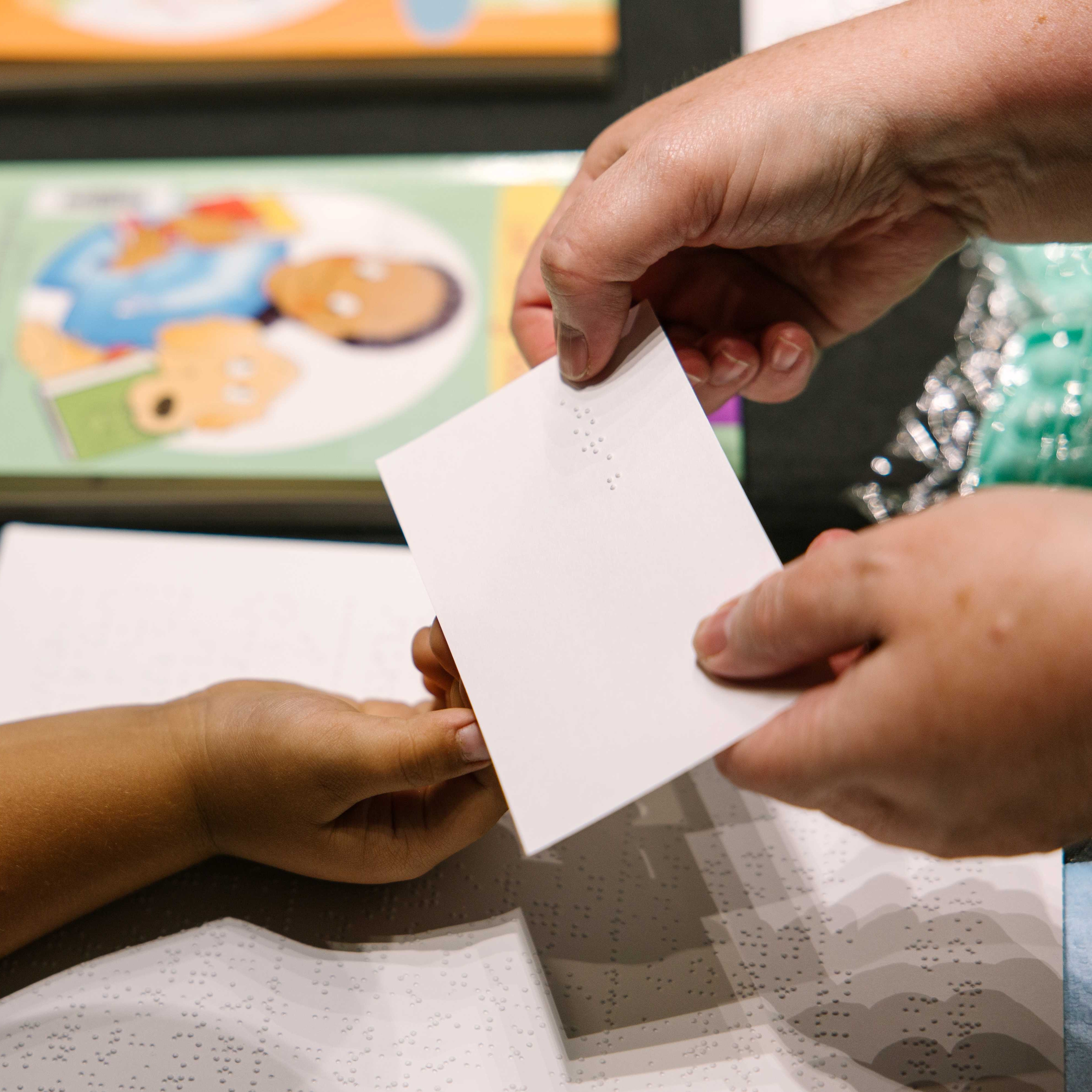 Two sets of hands, one adult handing a child a brailled notecard.